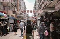 A busy street market bustling with activity, featuring various stalls and vendors selling clothing and other goods. Shoppers walk along the narrow pathway, with tall buildings lining each side. Signs with Chinese characters hang overhead, adding to the vibrant atmosphere.