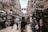 A busy street market bustling with activity, featuring various stalls and vendors selling clothing and other goods. Shoppers walk along the narrow pathway, with tall buildings lining each side. Signs with Chinese characters hang overhead, adding to the vibrant atmosphere.