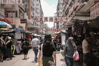 A busy street market bustling with activity, featuring various stalls and vendors selling clothing and other goods. Shoppers walk along the narrow pathway, with tall buildings lining each side. Signs with Chinese characters hang overhead, adding to the vibrant atmosphere.