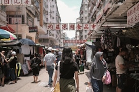 A busy street market bustling with activity, featuring various stalls and vendors selling clothing and other goods. Shoppers walk along the narrow pathway, with tall buildings lining each side. Signs with Chinese characters hang overhead, adding to the vibrant atmosphere.