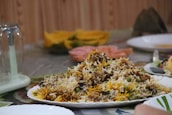 A plate filled with a colorful and aromatic rice dish, featuring long-grain rice mixed with vegetables and various spices. The platter is placed on a table set for dining, which includes other plates and a glass lid over a centerpiece. The background shows slices of fruit, adding a vibrant touch.