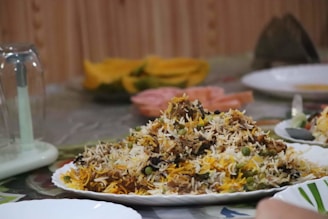 A plate filled with a colorful and aromatic rice dish, featuring long-grain rice mixed with vegetables and various spices. The platter is placed on a table set for dining, which includes other plates and a glass lid over a centerpiece. The background shows slices of fruit, adding a vibrant touch.