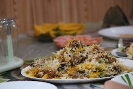 A plate filled with a colorful and aromatic rice dish, featuring long-grain rice mixed with vegetables and various spices. The platter is placed on a table set for dining, which includes other plates and a glass lid over a centerpiece. The background shows slices of fruit, adding a vibrant touch.