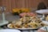 A plate filled with a colorful and aromatic rice dish, featuring long-grain rice mixed with vegetables and various spices. The platter is placed on a table set for dining, which includes other plates and a glass lid over a centerpiece. The background shows slices of fruit, adding a vibrant touch.