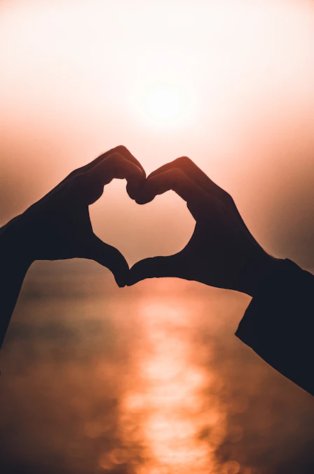 Two hands forming a heart shape silhouette against a warm glowing sky