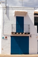 A white building with blue wooden shutters and door occupies the scene. A small balcony with a black iron railing is located above the large blue doors. The building has traditional Spanish architectural elements with a simple yet charming appearance. The shadows suggest strong sunlight, typical of a warm climate.