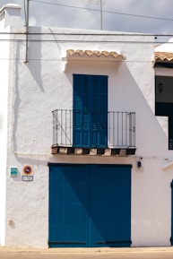 A white building with blue wooden shutters and door occupies the scene. A small balcony with a black iron railing is located above the large blue doors. The building has traditional Spanish architectural elements with a simple yet charming appearance. The shadows suggest strong sunlight, typical of a warm climate.