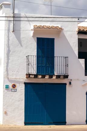 A white building with blue wooden shutters and door occupies the scene. A small balcony with a black iron railing is located above the large blue doors. The building has traditional Spanish architectural elements with a simple yet charming appearance. The shadows suggest strong sunlight, typical of a warm climate.