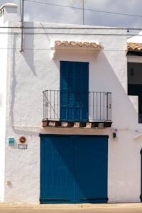 A white building with blue wooden shutters and door occupies the scene. A small balcony with a black iron railing is located above the large blue doors. The building has traditional Spanish architectural elements with a simple yet charming appearance. The shadows suggest strong sunlight, typical of a warm climate.