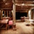 A rustic, open-air dining area with wooden tables and chairs, some with red cushions. The space features a high wooden ceiling with exposed beams, contributing to a warm and inviting atmosphere. Soft lighting adds coziness to the setting, with large windows providing potential views outside.
