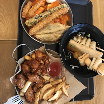 A table with a variety of Korean street food, including tteokbokki with fish cakes and crispy fried toppings, chicken skewers, and a bowl of oden with skewered fish cakes in broth. There are also fried potato wedges and a container of sauce.