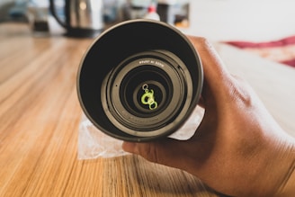 A hand holding a camera lens on a wooden surface. The lens features visible markings inside, indicating it is made in Japan. The background is slightly blurred, suggesting a shallow depth of field.