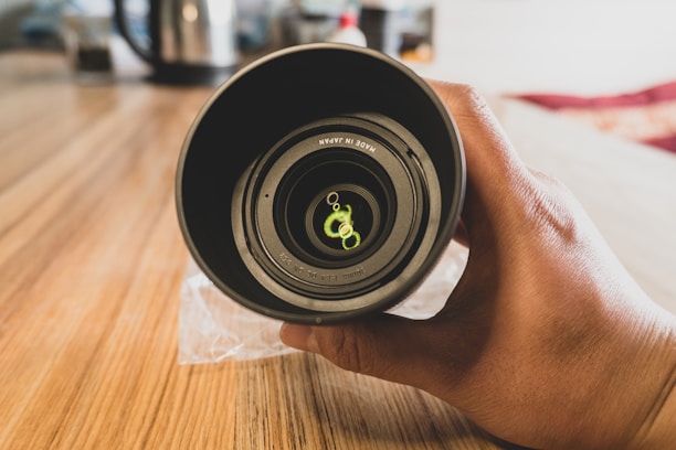 A hand holding a camera lens on a wooden surface. The lens features visible markings inside, indicating it is made in Japan. The background is slightly blurred, suggesting a shallow depth of field.