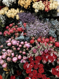 A vibrant assortment of various flowers, including red roses, pink peonies, yellow chrysanthemums, lavender sprigs, and white roses, all arranged in a display. The flowers are fresh and blooming, creating a lush and colorful scene with green foliage accentuating the mix.