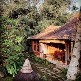 A traditional wooden house with a shaded porch is surrounded by lush greenery. The architecture includes a terracotta-tiled roof and wooden furniture on the porch, partially lit by sunlight. Stone paths lead through a garden with abundant tropical plants.
