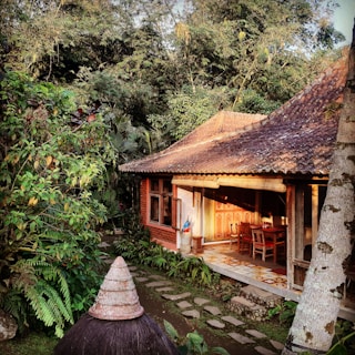 A traditional wooden house with a shaded porch is surrounded by lush greenery. The architecture includes a terracotta-tiled roof and wooden furniture on the porch, partially lit by sunlight. Stone paths lead through a garden with abundant tropical plants.