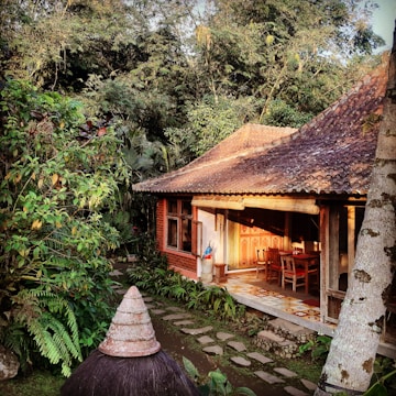 A traditional wooden house with a shaded porch is surrounded by lush greenery. The architecture includes a terracotta-tiled roof and wooden furniture on the porch, partially lit by sunlight. Stone paths lead through a garden with abundant tropical plants.