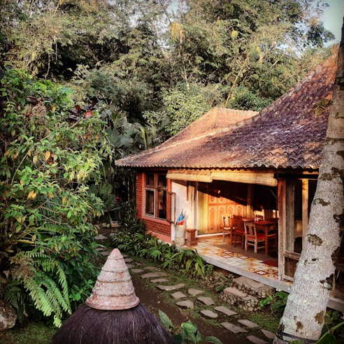 A traditional wooden house with a shaded porch is surrounded by lush greenery. The architecture includes a terracotta-tiled roof and wooden furniture on the porch, partially lit by sunlight. Stone paths lead through a garden with abundant tropical plants.