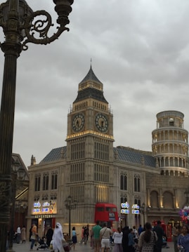 The image features a replica of the iconic Big Ben tower and the Leaning Tower of Pisa, surrounded by a lively setting with shops and people walking around. A red double-decker bus is parked in front, and colorful storefront signs are visible. The sky is overcast, adding a muted atmosphere to the scene.