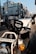 A close-up view of a Honda CC50 motorcycle parked on an urban street. The image focuses on the front part of the motorcycle, highlighting its headlamp, handlebar, and brand logo. In the background, there are modern apartment buildings and some parked vehicles, suggesting a city setting.