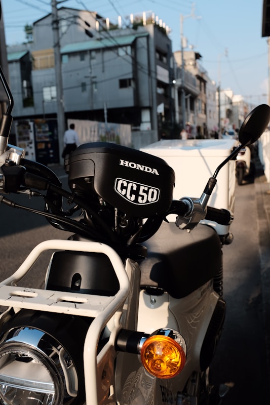 A close-up view of a Honda CC50 motorcycle parked on an urban street. The image focuses on the front part of the motorcycle, highlighting its headlamp, handlebar, and brand logo. In the background, there are modern apartment buildings and some parked vehicles, suggesting a city setting.