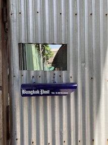 A corrugated metal wall with a rectangular mesh-covered opening allowing a partial view of trees and a building outside. A blue newspaper holder with the text 'Bangkok Post' and a contact number is attached to the wall.
