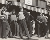A group of people stand outside a building under a striped awning. The group consists mostly of men, with one woman visible. The men are dressed in casual, working-class clothing typical of the early 20th century. One man in the foreground is posing with his hand on his hip, looking confident and possibly engaged in a conversation or speech. The sign above reads 'Organized Labor,' suggesting the gathering is related to labor rights or union activity.