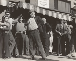 A group of people stand outside a building under a striped awning. The group consists mostly of men, with one woman visible. The men are dressed in casual, working-class clothing typical of the early 20th century. One man in the foreground is posing with his hand on his hip, looking confident and possibly engaged in a conversation or speech. The sign above reads 'Organized Labor,' suggesting the gathering is related to labor rights or union activity.