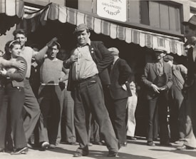 A group of people stand outside a building under a striped awning. The group consists mostly of men, with one woman visible. The men are dressed in casual, working-class clothing typical of the early 20th century. One man in the foreground is posing with his hand on his hip, looking confident and possibly engaged in a conversation or speech. The sign above reads 'Organized Labor,' suggesting the gathering is related to labor rights or union activity.