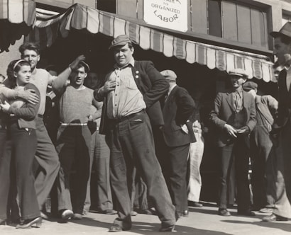 A group of people stand outside a building under a striped awning. The group consists mostly of men, with one woman visible. The men are dressed in casual, working-class clothing typical of the early 20th century. One man in the foreground is posing with his hand on his hip, looking confident and possibly engaged in a conversation or speech. The sign above reads 'Organized Labor,' suggesting the gathering is related to labor rights or union activity.