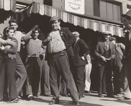 A group of people stand outside a building under a striped awning. The group consists mostly of men, with one woman visible. The men are dressed in casual, working-class clothing typical of the early 20th century. One man in the foreground is posing with his hand on his hip, looking confident and possibly engaged in a conversation or speech. The sign above reads 'Organized Labor,' suggesting the gathering is related to labor rights or union activity.