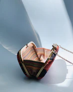 A small, woven handbag with multicolored horizontal stripes in a variety of vibrant hues. The bag features gold-tone metal edges and includes a delicate metallic chain strap. It is set against a smooth, light blue background with soft shadows cast across the surface.