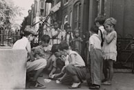 A group of children are gathered on a sidewalk, with some sitting and others standing. The children are engaged in a card game, and the setting appears to be in an urban residential area with brick buildings and a wrought iron fence in the background. The children wear casual clothing typical of the mid-20th century.