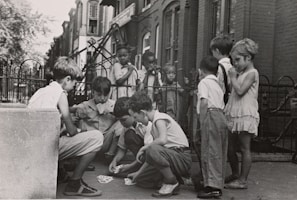 A group of children are gathered on a sidewalk, with some sitting and others standing. The children are engaged in a card game, and the setting appears to be in an urban residential area with brick buildings and a wrought iron fence in the background. The children wear casual clothing typical of the mid-20th century.