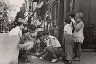 A group of children are gathered on a sidewalk, with some sitting and others standing. The children are engaged in a card game, and the setting appears to be in an urban residential area with brick buildings and a wrought iron fence in the background. The children wear casual clothing typical of the mid-20th century.