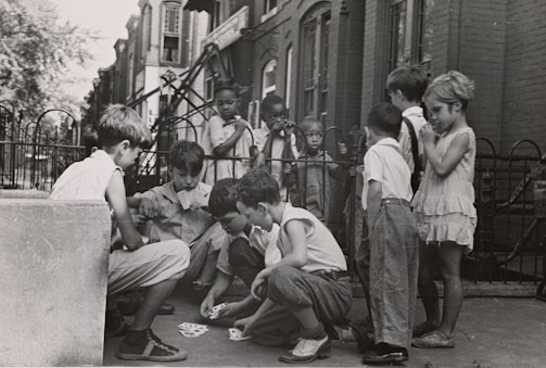 A group of children are gathered on a sidewalk, with some sitting and others standing. The children are engaged in a card game, and the setting appears to be in an urban residential area with brick buildings and a wrought iron fence in the background. The children wear casual clothing typical of the mid-20th century.
