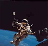 An astronaut is floating in the vastness of space with the Earth visible in the background. Connected to equipment by a gold-hued tether, the astronaut's suit reflects some light, indicating the sun's presence nearby. The darkness of space contrasts sharply with the blue curvature of the Earth below.