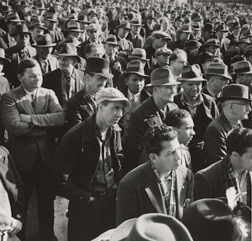 A large group of men stands closely together, most wearing hats and suits, suggesting a formal or organized gathering. The men appear focused and serious.