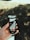 A hand is holding a can of Nescafé Xpress Black Roast, featuring a sleek black design with coffee bean graphics. The background is a blurred outdoor setting with dappled sunlight filtering through trees.