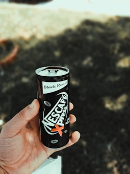 A hand is holding a can of Nescafé Xpress Black Roast, featuring a sleek black design with coffee bean graphics. The background is a blurred outdoor setting with dappled sunlight filtering through trees.