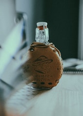 A decorative glass bottle with a ceramic cover sits on a wooden surface. The cover features an embossed image of a person wearing a hat and smoking, along with text in a foreign language. The bottle is topped with a swing stopper. The background is softly blurred, indicating an indoor setting.