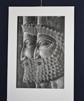 A black and white engraving or relief showing two male figures in profile, wearing intricately detailed headdresses and beards. The artwork is reminiscent of ancient styles, with precise and ornate curls in the hair and beard.