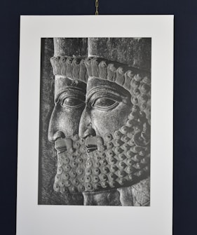 A black and white engraving or relief showing two male figures in profile, wearing intricately detailed headdresses and beards. The artwork is reminiscent of ancient styles, with precise and ornate curls in the hair and beard.