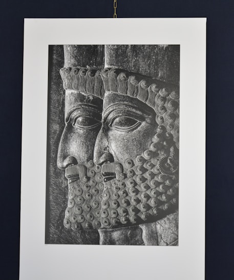 A black and white engraving or relief showing two male figures in profile, wearing intricately detailed headdresses and beards. The artwork is reminiscent of ancient styles, with precise and ornate curls in the hair and beard.