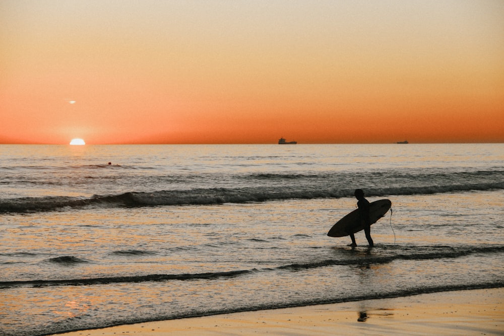 silhouette of person with surfboard in beach at sunset