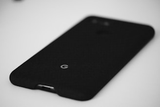 A black phone case with a fabric texture is placed on a plain white surface. The case displays a small, centered logo near the bottom.
