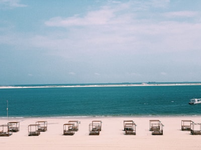 An empty beach with a row of wooden cabanas or sun loungers evenly spaced on the sand. The calm blue ocean extends to the horizon, with a small boat visible to the right. A sandbar or distant shoreline is visible further out.