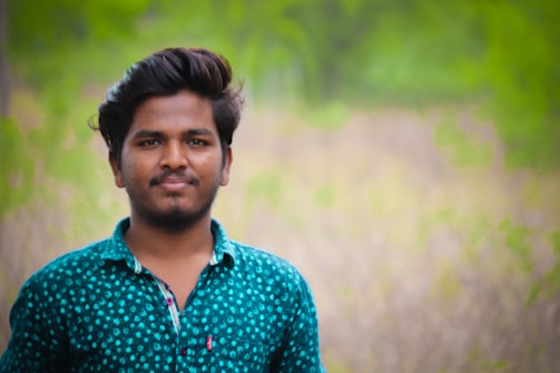 A person with dark hair and a beard wears a teal shirt with white patterns, standing in front of a blurred natural background. The setting appears lush and green, suggesting a park or forest area.