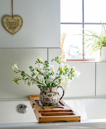 A bathroom scene with a light-filled window and a wall-mounted heart-shaped decoration with the words 'happy hearts are filled with love.' A floral-patterned jug filled with white flowers rests on a bamboo bathtub tray. Next to it, a potted plant adds greenery to the window ledge.