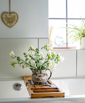 A bathroom scene with a light-filled window and a wall-mounted heart-shaped decoration with the words 'happy hearts are filled with love.' A floral-patterned jug filled with white flowers rests on a bamboo bathtub tray. Next to it, a potted plant adds greenery to the window ledge.